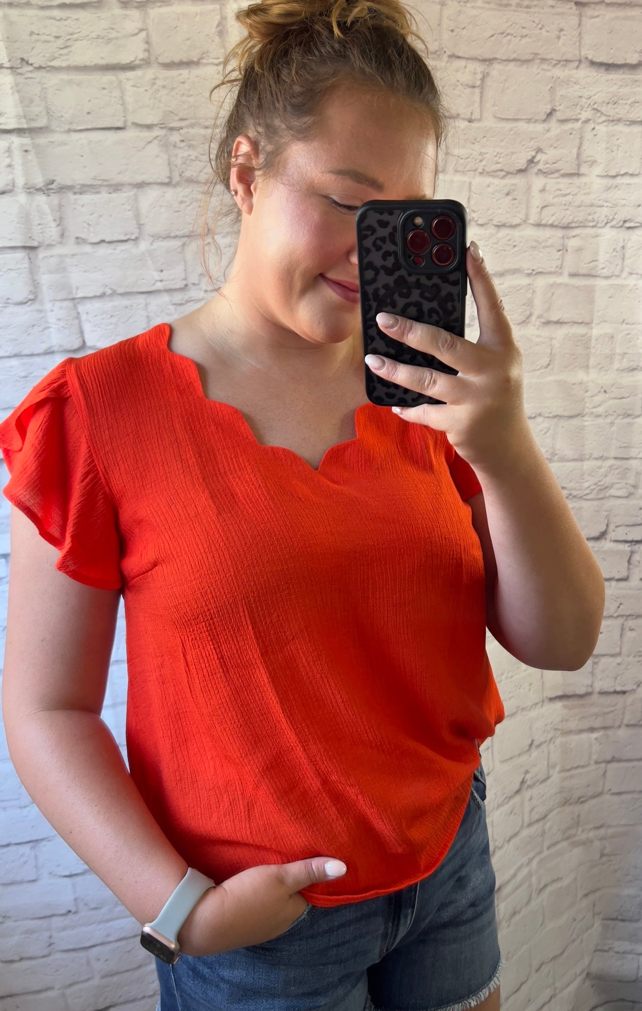 Person wearing an orange top and blue shorts taking a mirror selfie against a brick wall.