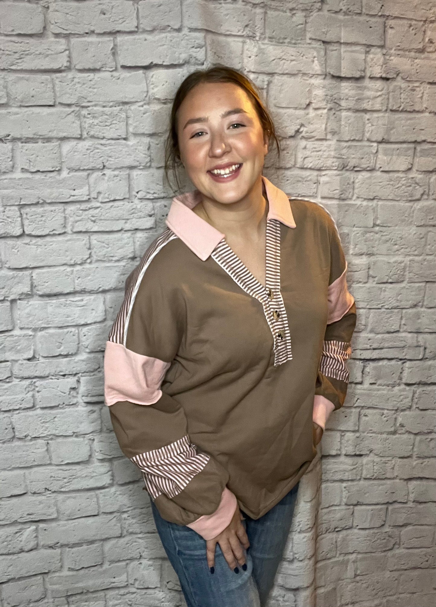 A woman is wearing a brown pullover featuring a pink collar, pink sleeve accents, and striped brown-and-white detailing along the neckline and sleeves.