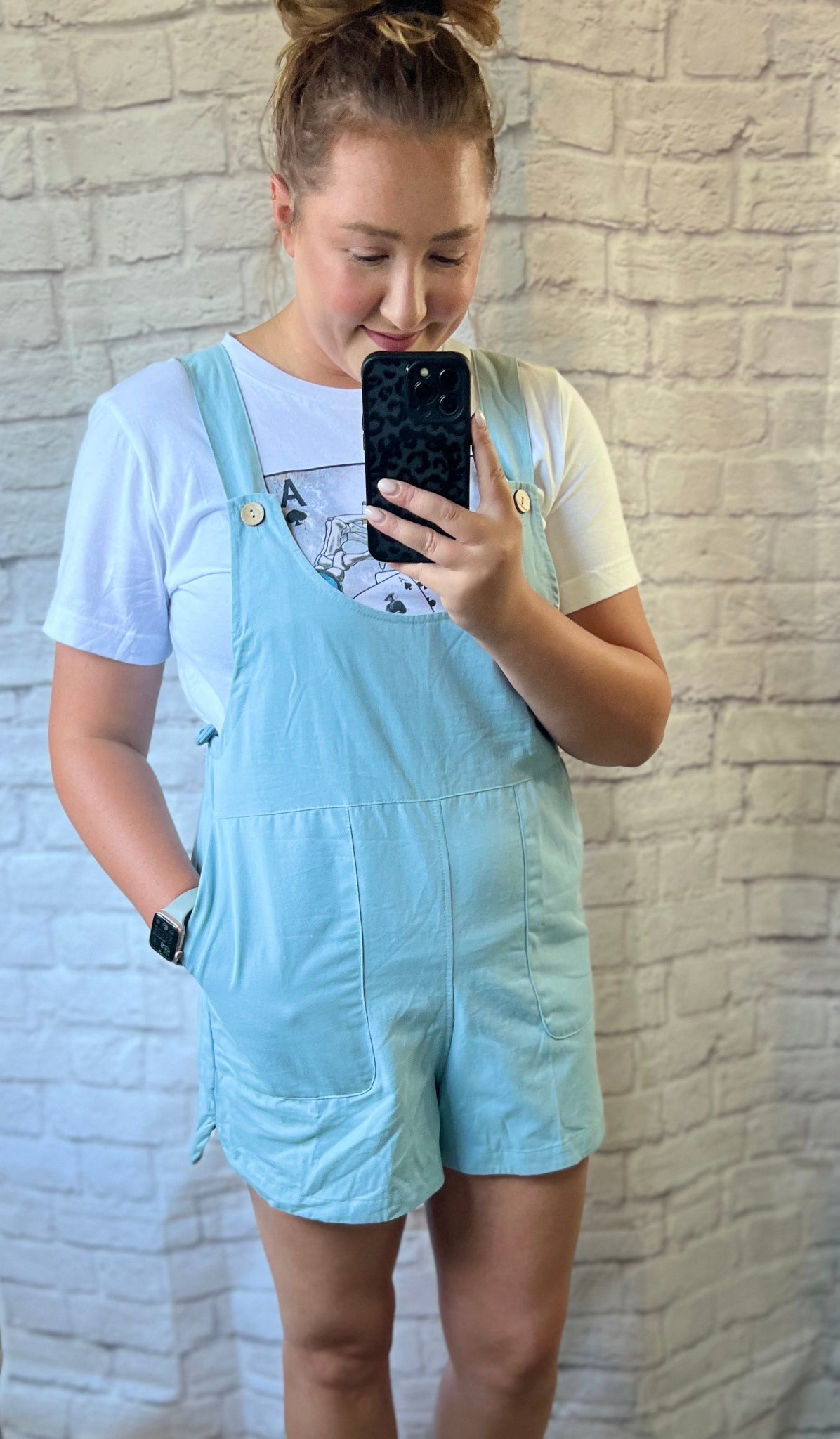 Person wearing light blue overalls taking a mirror selfie against a white brick wall.