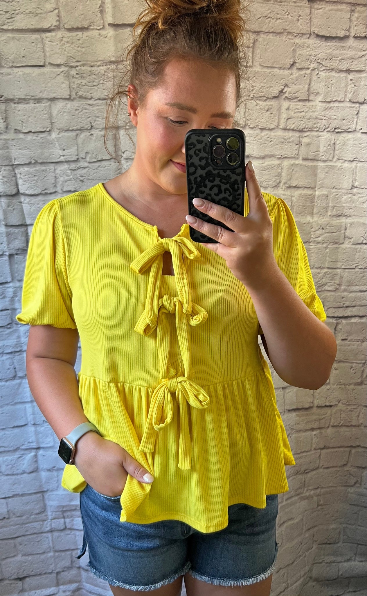 Person wearing a yellow top with ruffled details, taking a mirror selfie against a brick wall.