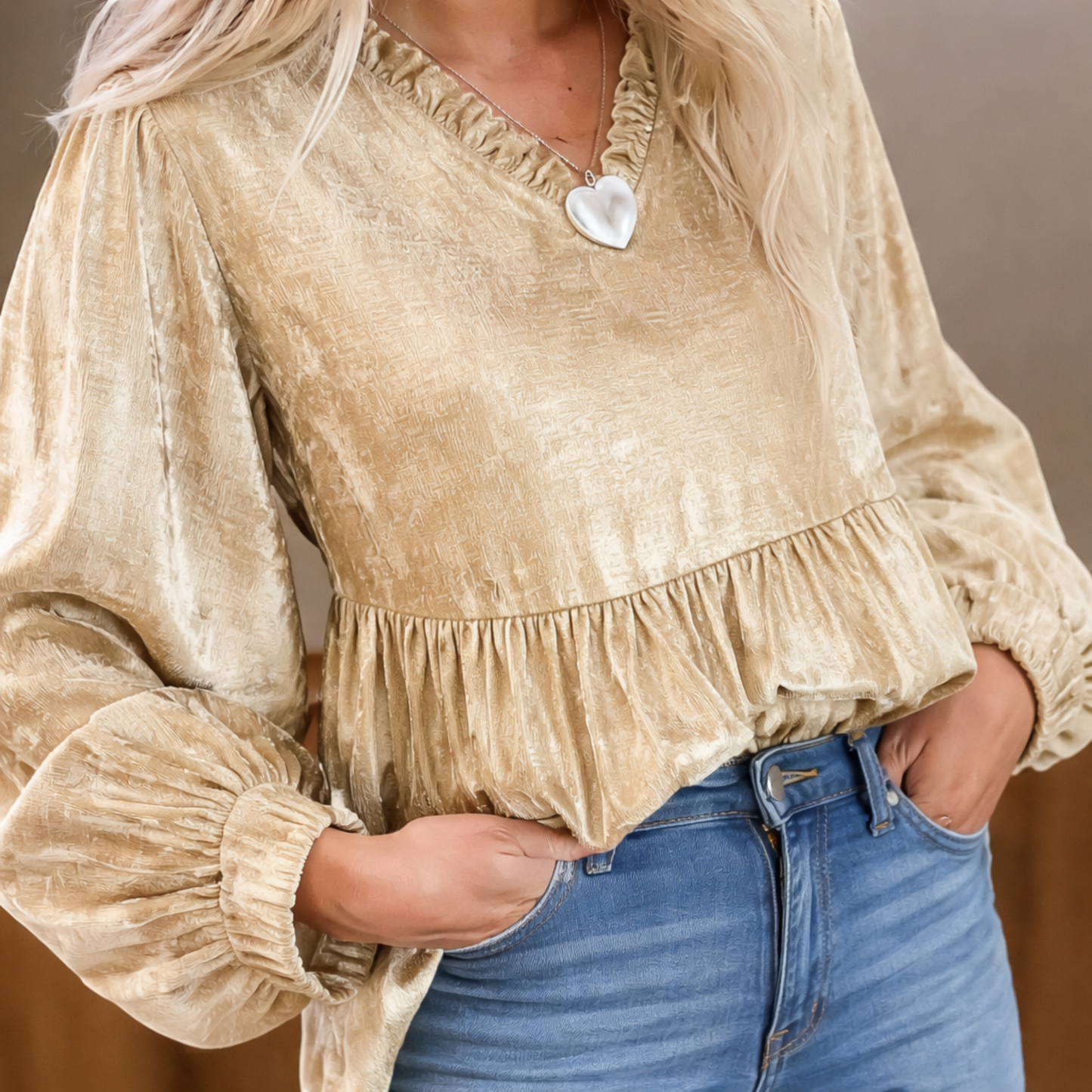 Person wearing a beige blouse with ruffled details and blue jeans against a neutral background