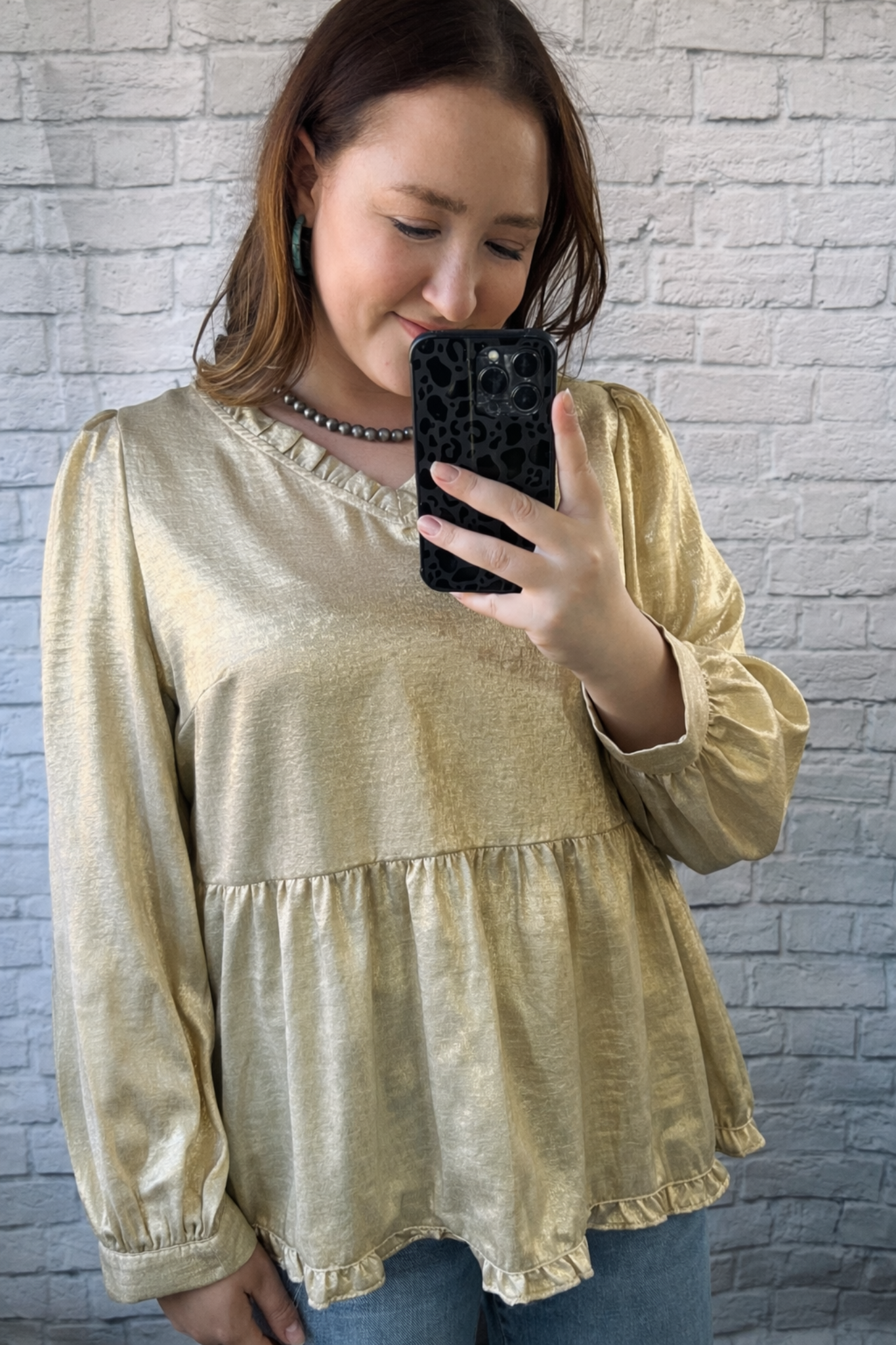 Woman taking a mirror selfie wearing a gold blouse against a gray brick wall.