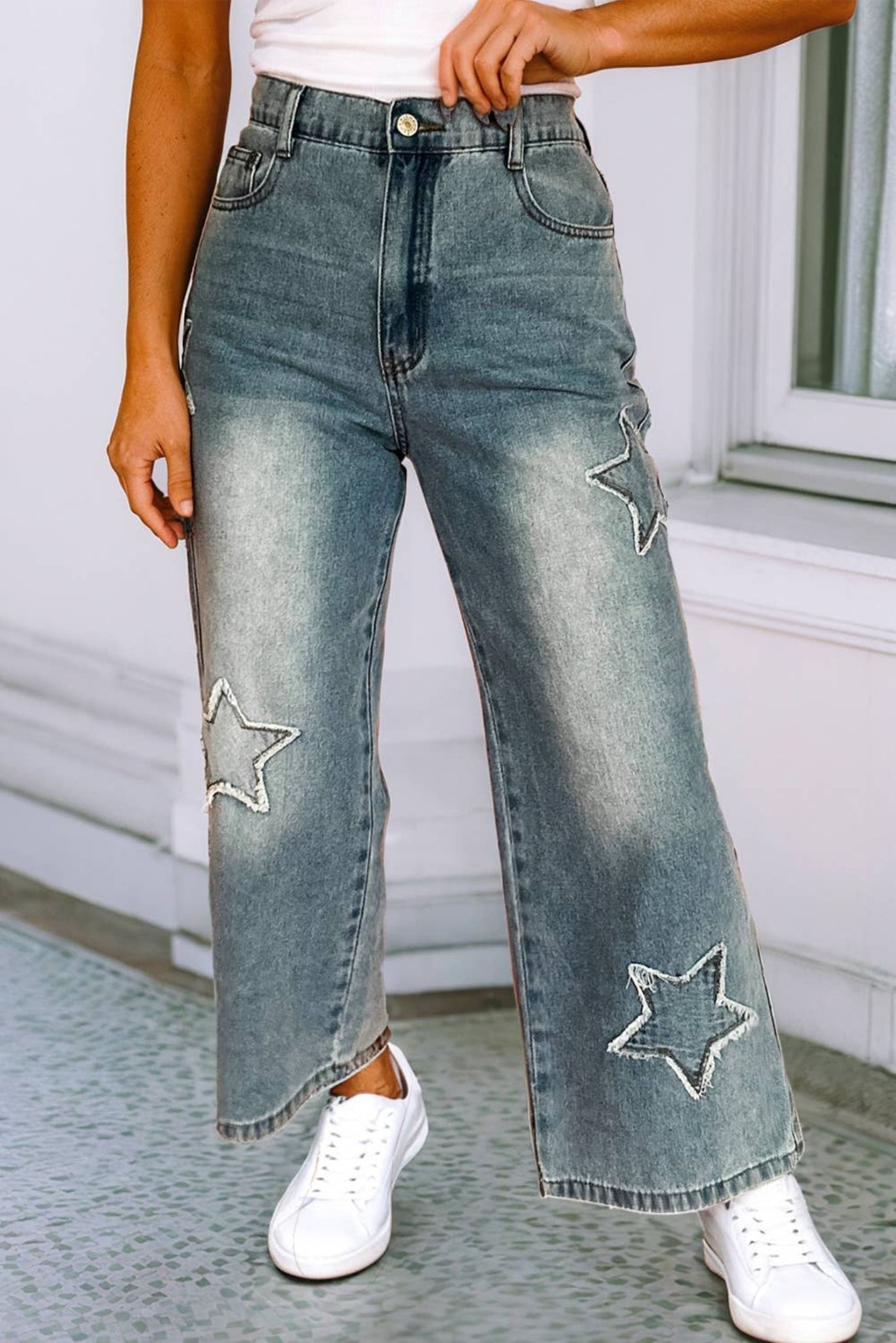 A pair of light wash wide leg high waist jeans with star patchwork details on the knees, paired with a white top and white sneakers.