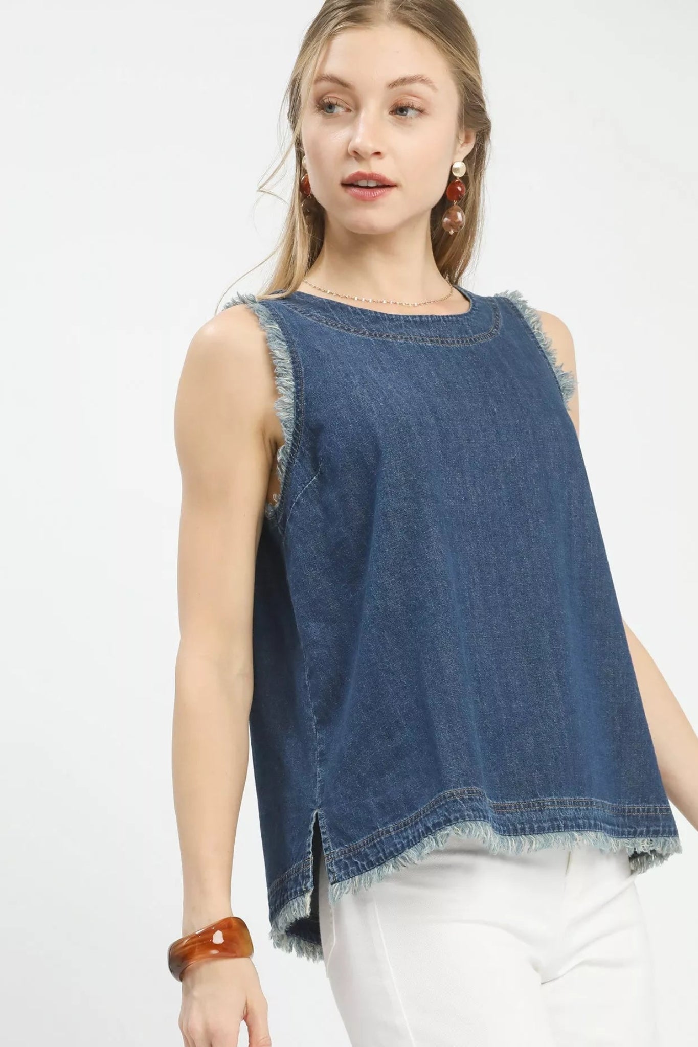 Woman wearing a sleeveless denim tank top with frayed trim edges and a relaxed fit, styled with white pants for a casual boutique look.