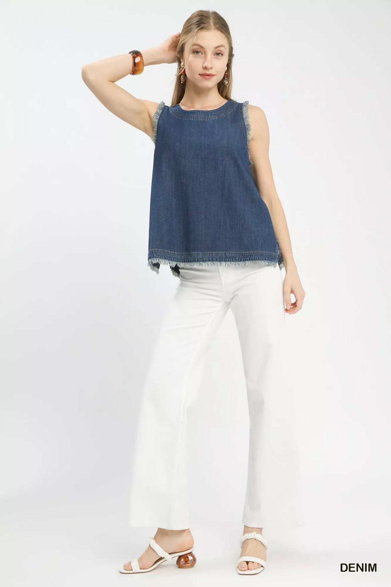 Woman wearing a sleeveless denim tank top with frayed trim edges and a relaxed fit, styled with white pants for a casual boutique look.