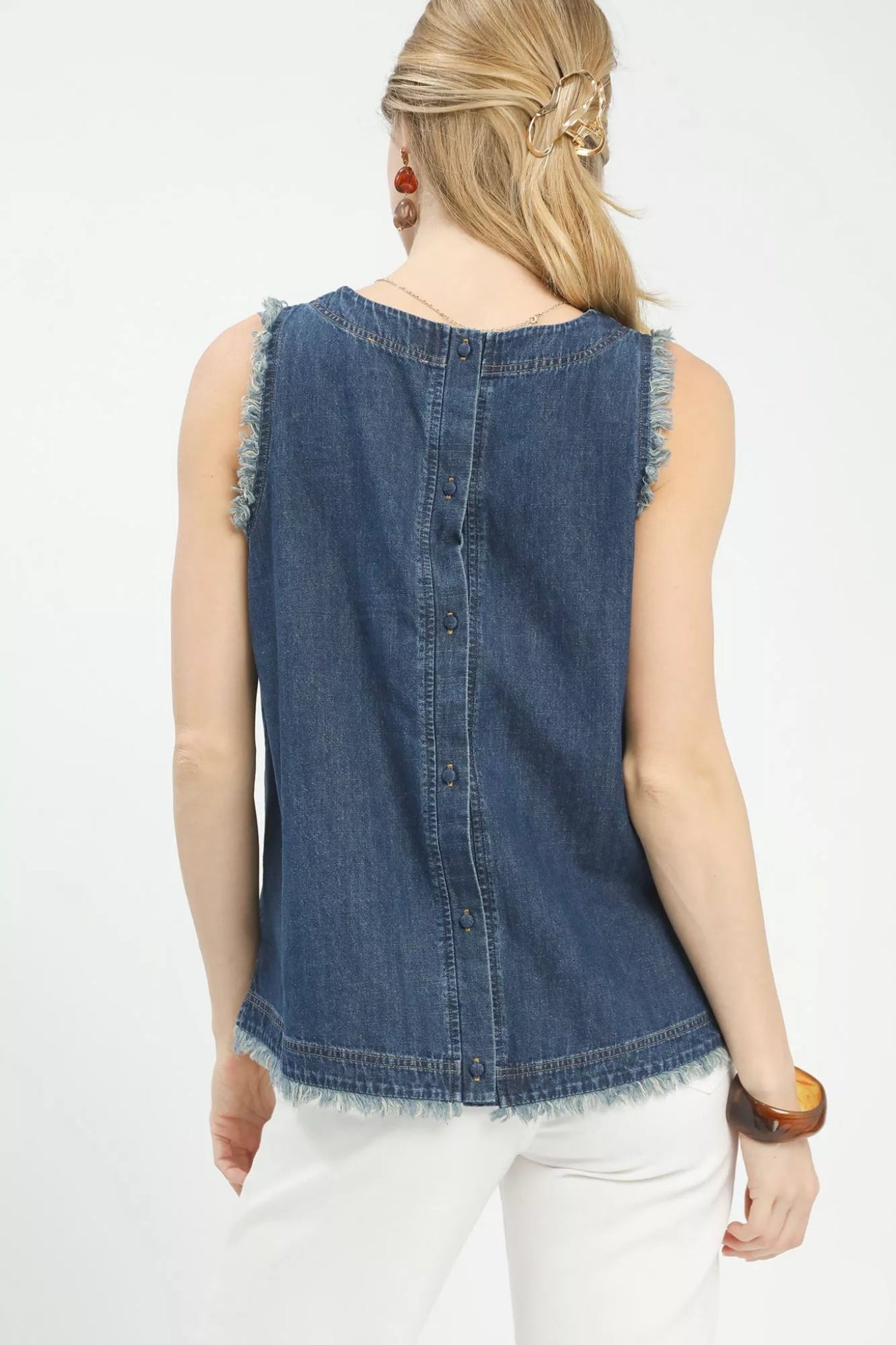 Woman wearing a sleeveless denim tank top with frayed trim edges and a relaxed fit, styled with white pants for a casual boutique look.