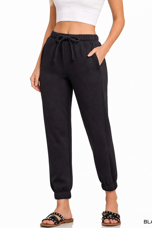 Model wearing black jogger sweatpants with elastic waistband, drawstring tie, cuffed ankles, and white cropped tank top.