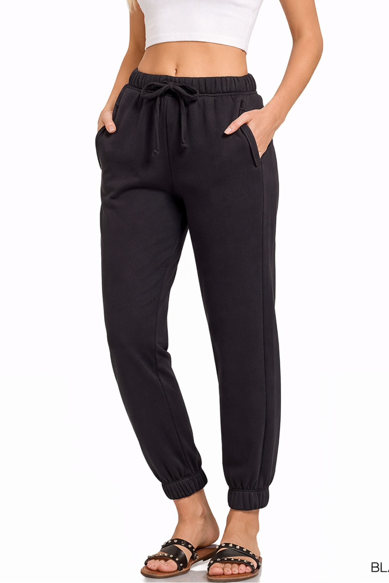 Model wearing black jogger sweatpants with elastic waistband, drawstring tie, cuffed ankles, and white cropped tank top.