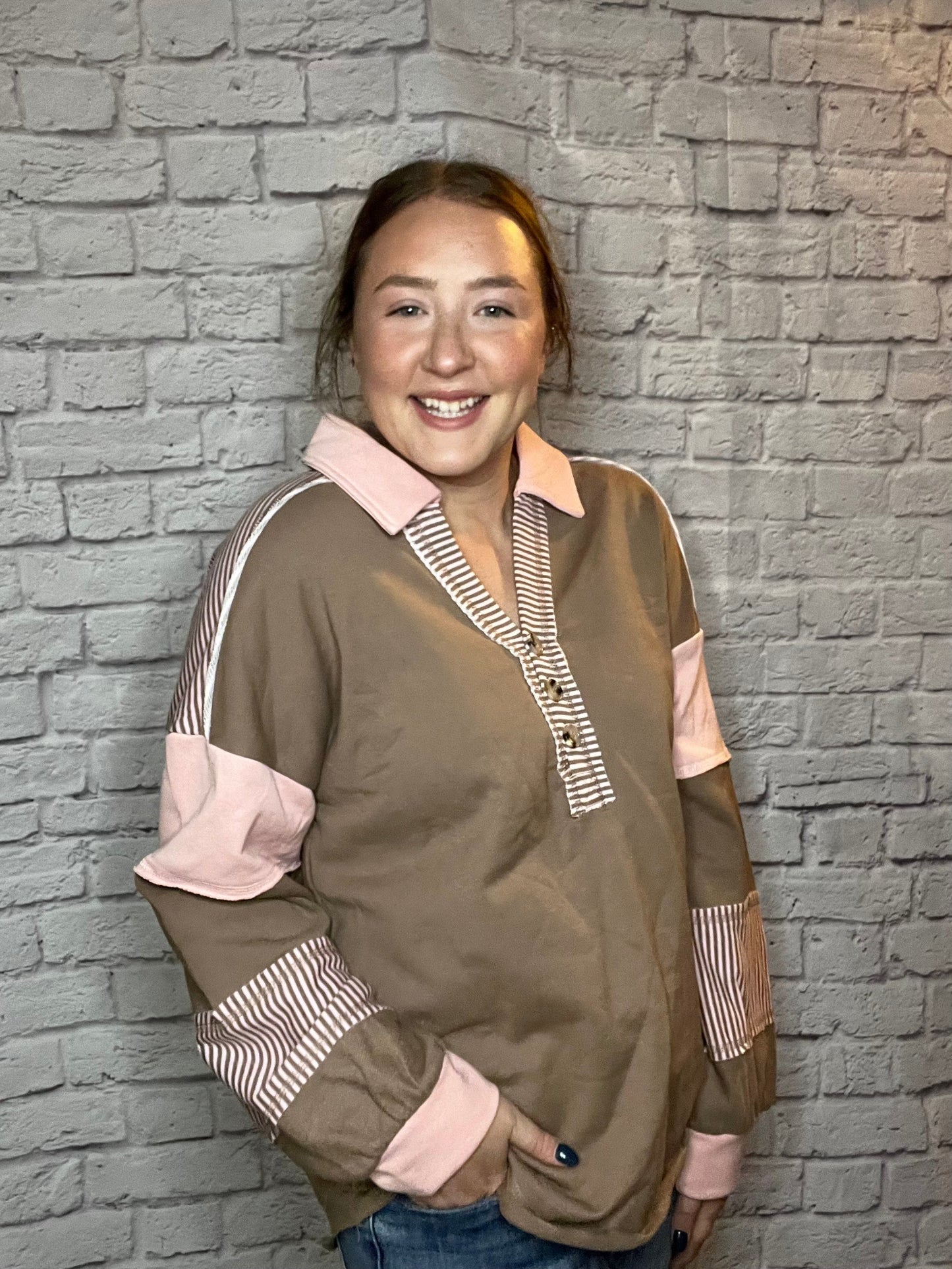 A woman is wearing a brown pullover featuring a pink collar, pink sleeve accents, and striped brown-and-white detailing along the neckline and sleeves.