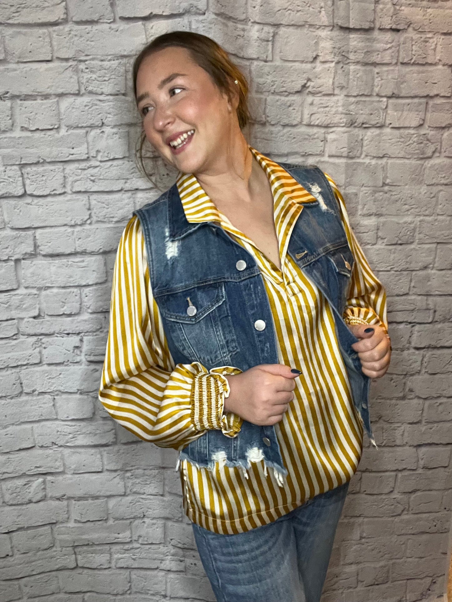 A woman wearing a mustard yellow and white striped blouse layered with a distressed blue denim vest.