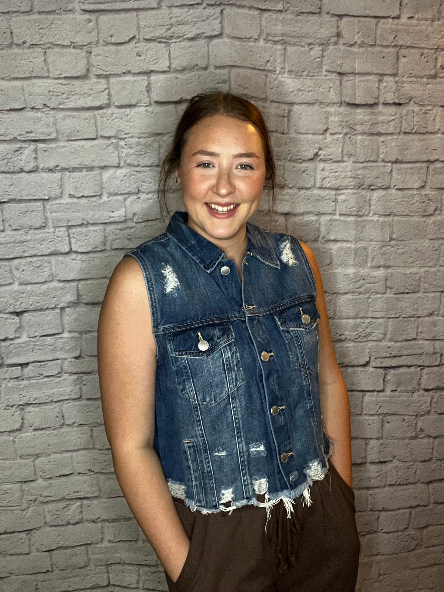 A woman wearing a sleeveless distressed denim vest with frayed hem and front button closure.