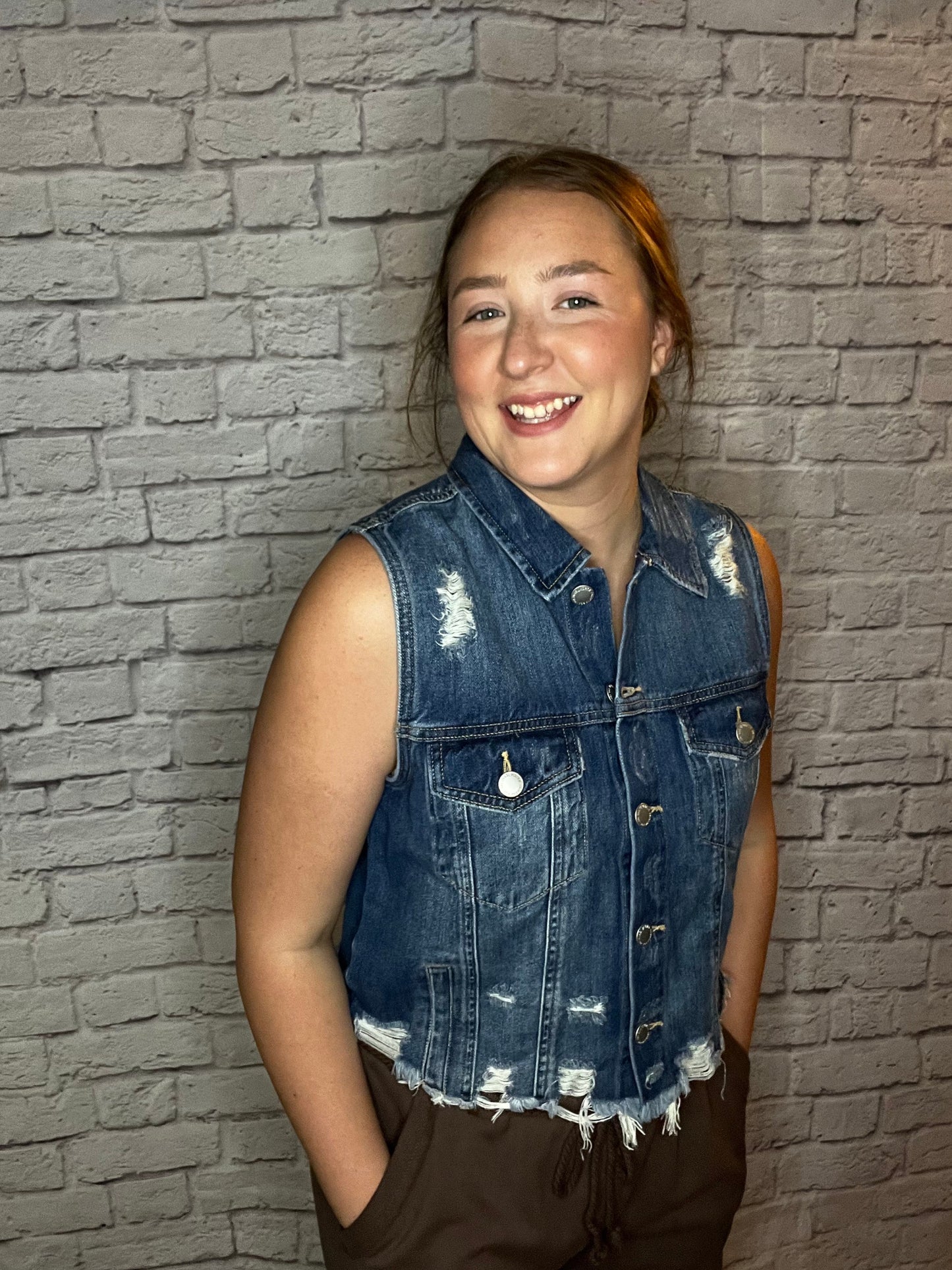 A woman wearing a sleeveless distressed denim vest with frayed hem and front button closure.