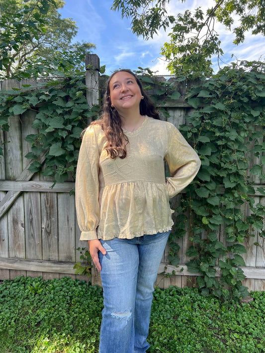 Woman taking a mirror selfie wearing a gold blouse against a gray brick wall.