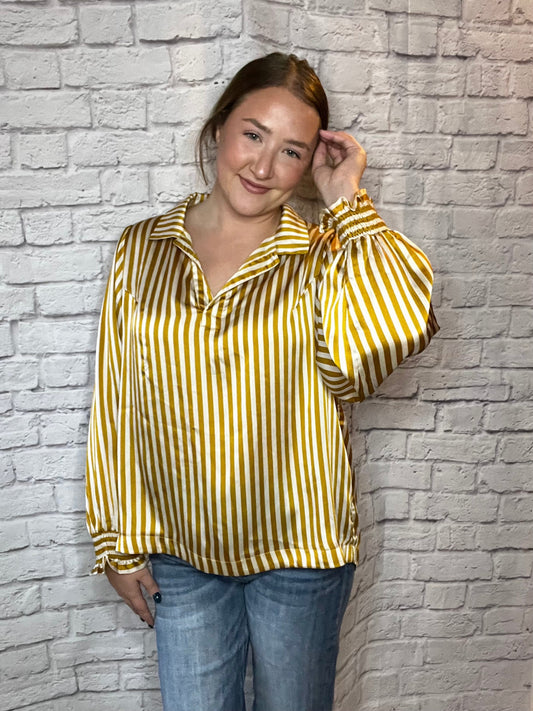 A woman wearing a mustard yellow and white vertical striped blouse with a relaxed fit, collared neckline, and gathered sleeves.